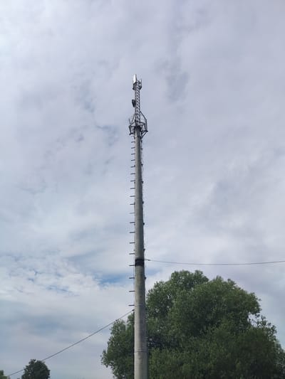 A tall concrete utility pole, topped with a metal communication tower, stands prominently against a cloudy sky. The tower features multiple antennas and a ladder running up its side. In the foreground, the lush green foliage of large trees frames the base of the pole. The scene appears to be set in an outdoor, possibly rural or suburban area, with a hint of a power line stretching across the frame. The weather suggests a partly cloudy day. This image was captured in the city of Trestna, Russia.