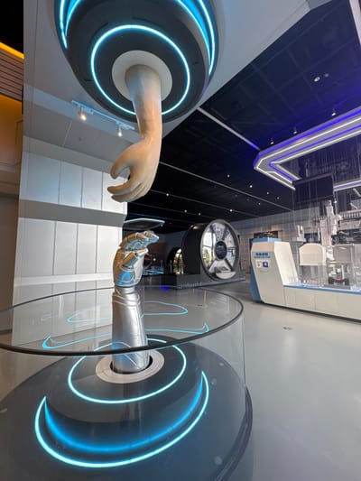 This image displays a modern, technologically themed exhibition hall located in Guangming District, China. The central elements are two large hand sculptures: a beige, human-like hand extending downwards from a black ceiling fixture illuminated with circular blue neon lights, and a silver robotic forearm and hand extending upwards from a circular glass display table. The base of the display table features concentric blue neon lighting. In the background, a large, dark circular display unit with segmented panels and text is visible to the left. To the right, a white and light blue exhibit counter presents items within clear rectangular displays, indicative of laboratory equipment or technological components, with a Chinese-language sign attached. The floor is a light grey, and the dark ceiling incorporates recessed lighting and linear blue and white strip lighting, creating a futuristic ambiance. No individuals are present.