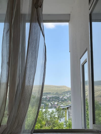 A window view features a sheer curtain draped across the lower portion of the frame. Outside, a clear blue sky is visible above a landscape with a tree and some houses. The scene seems to be reflected in the window glass. The architecture surrounding the window includes a white ceiling and frame, with a smaller rectangular window set into the ceiling.