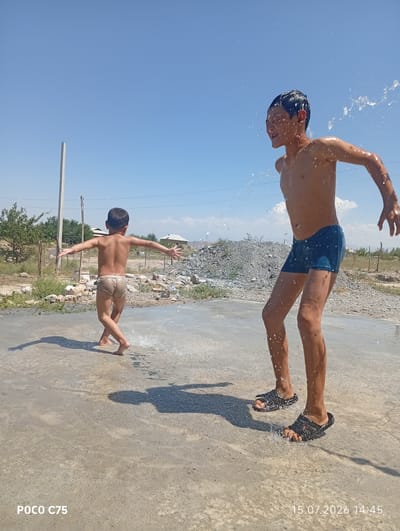 Two young boys are playing in shallow water outdoors on a sunny day. The boy on the right, wearing blue swimming trunks and black sandals, is looking to his left with water splashing around him. He has his arms extended outwards. The boy on the left, wearing light-colored briefs, is facing away from the camera with his arms outstretched to the sides. He appears to be mid-twirl, with water splashing around his feet. The scene is set on a paved surface that is wet and reflective. In the background, there are some sparse trees, rocks, and distant buildings. The sky is clear and blue. The time stamp on the image indicates July 15, 2026, at 14:45. The location context is Чоң-Гара, Kyrgyzstan. The overall mood is joyful and playful.