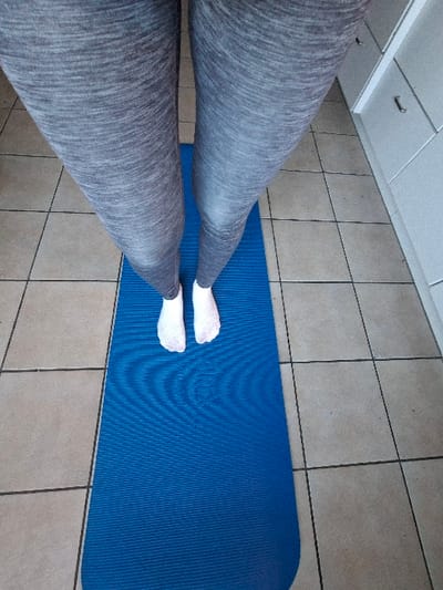The scene depicts a high-angle view of an individual's lower body within an indoor environment, stated to be in Davos, Switzerland. The person is wearing grey heathered leggings and white socks. They stand with their feet positioned towards the upper end of a royal blue, textured exercise mat. The mat rests on a floor comprised of light beige square tiles with dark grout lines. To the right of the mat, in the background, a light-colored cabinet or set of drawers is visible, featuring a dark, curved handle on one drawer. The overall environment suggests a domestic setting, likely a kitchen or utility room. No explicit actions beyond standing are discernible.