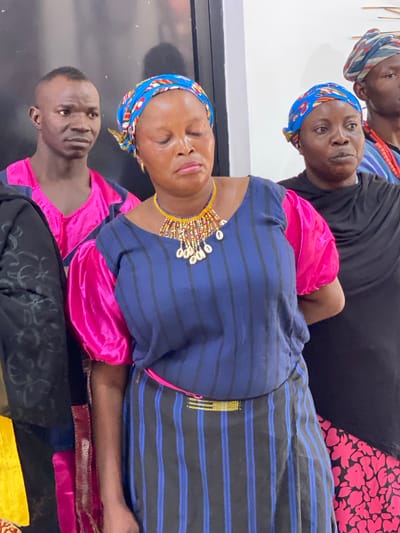 This image captures a group of people, likely women and one man, in what appears to be an indoor setting in Kwarau, Nigeria. The lighting suggests it could be daytime indoors or under artificial light.
In the foreground, a woman wears a blue and yellow patterned headscarf and a navy blue top with thin blue horizontal stripes. She also wears a prominent, multi-colored beaded necklace adorned with white cowrie shells around her neck. Her eyes are closed, and her lips are slightly parted, giving her a serene or contemplative expression. Beneath her blue top, a vibrant pink garment is visible, possibly part of her sleeves or an underlayer.
Behind her, to her left, another woman is visible. She wears a blue headscarf with red and yellow patterns, a black top, and an orange beaded necklace. She is looking directly at the camera with a neutral expression. Further back and to the top left, a third woman is partially seen, wearing a striped headscarf (blue, red, and white) and a black top, also looking towards the viewer.
On the bottom left, partially cropped, a man with a short haircut is visible, wearing a pink garment under a dark top, also looking towards the camera with a neutral expression. The subjects' attire, including patterned headscarves and beaded jewelry, suggests cultural or traditional clothing common in the region. The background consists of plain dark and light walls, providing a simple backdrop for the group. There is no visible text in the image.