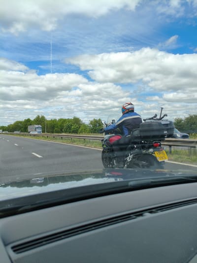 The image shows a perspective from inside a vehicle looking out onto a motorway, likely the A500, heading towards Newcastle-under-Lyme, United Kingdom. A motorcycle with a rider wearing a blue and black jacket and a white, blue, and orange helmet is in the foreground, to the right of the lane. The motorcycle has a large black storage box on the back and a yellow license plate with the characters "YP15 YKS." A blue car is visible behind the motorcycle. The road ahead shows lane markings and a safety barrier. On the left side of the road, a large articulated lorry with "E.V.T.S." written on its side is passing. Beyond the road, a line of dense green trees forms the horizon under a partly cloudy blue sky. The reflection of the road, trees, and sky is visible on the dashboard of the vehicle from which the photograph is taken.