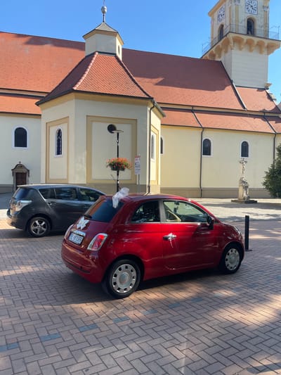 In Stupava, Slovakia, in front of a church, a red Fiat 500 adorned with a white bow is parked on a brick-paved area. A dark gray hatchback is parked behind it. The church features a terracotta tiled roof and a tall clock tower. A stone cross stands in front of the church, with trees and a clear blue sky in the background.