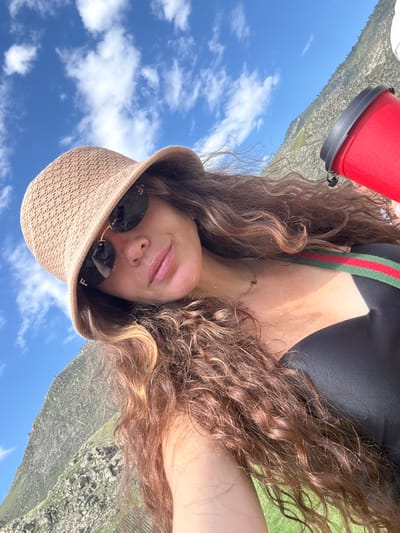 A woman is taking a selfie outdoors in Kupchegen, Russia. She is wearing a straw bucket hat and sunglasses, and has long, curly brown hair. She is also wearing a black swimsuit with a red and green striped strap. A red thermos is visible in the upper right corner of the image. In the background, a steep, rocky hillside covered in sparse green vegetation slopes upwards against a bright blue sky with scattered white clouds. The sun is shining, casting shadows on the woman's face and neck.