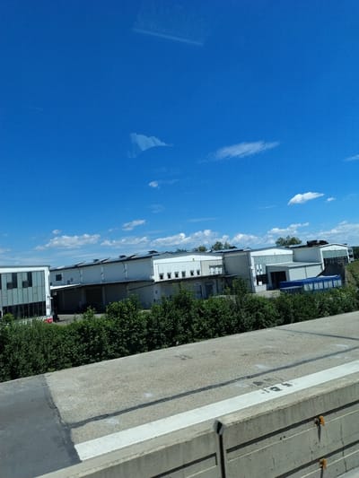 A wide shot captures a bright, sunny day with a clear blue sky dotted with a few white clouds. In the foreground, a concrete barrier with visible horizontal lines and some rust-colored attachments runs across the bottom of the frame. Behind the barrier, a grey asphalt road with black markings and a white painted line is visible.
In the mid-ground, a row of modern industrial buildings with white facades and dark-tinted windows stretches across the scene. The roofs of these buildings are equipped with visible solar panels, and several large industrial doors or loading bays are apparent. Adjacent to the buildings, dense green trees and bushes form a thick line of vegetation. One of the buildings has a more modern facade with a grid of dark windows.
In the background, more trees and buildings are visible, suggesting an industrial area within or on the outskirts of the city of Hengersberg, Germany. The overall impression is one of an organized and functional industrial landscape under a clear sky.
