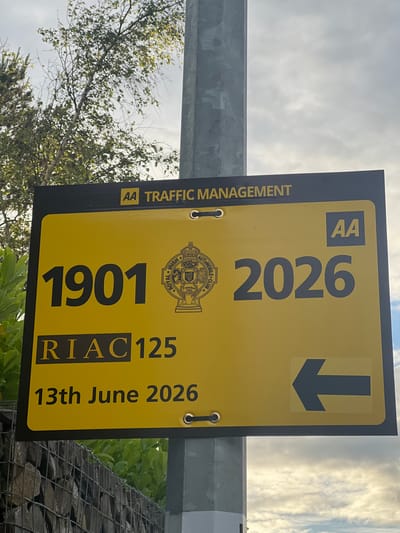 The image displays a yellow sign attached to a metal pole. The sign has black text and graphics. At the top, "AA TRAFFIC MANAGEMENT" is printed in white, with "AA" also appearing in yellow on the right side. Below this, the numbers "1901" are displayed prominently on the left, and "2026" on the right. Between these numbers is a circular emblem with a crown on top, containing text that includes "ROYAL IRISH AUTOMOBILE CLUB" and "FOUNDED 1901". Below the numbers, "RIAC 125" is written, followed by "13th June 2026". On the right side of the sign, a black arrow points to the left. The background consists of a cloudy sky and foliage. The sign appears to be in an outdoor setting, possibly near a road or a street. The mention of "Balbriggan, Ireland" is not supported by any visual evidence within the image itself; this information appears to be external context.