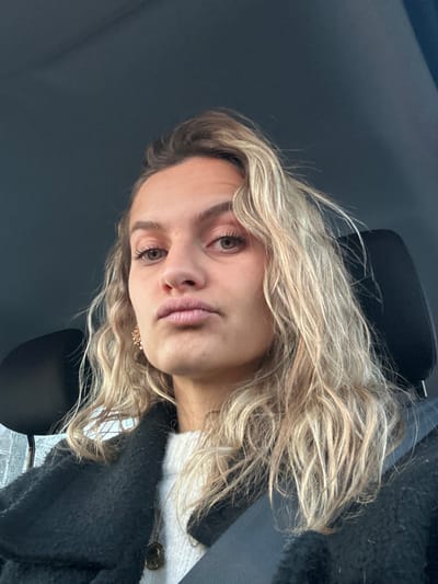 A young woman with wavy blonde hair, hazel eyes, and full lips poses in what appears to be a car. She wears a dark fuzzy cardigan over a white sweater with large buttons, a gold earring visible on one ear. A black headrest is behind her and a seatbelt crosses her chest. The car's interior is a dark gray. The woman's expression is neutral.