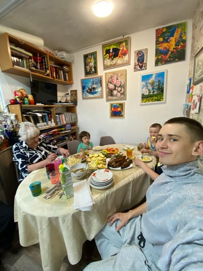 The image shows a domestic interior, seemingly in a home in Тушна, Russia, judging by the visible objects and style. Several individuals are present at a table laden with food and tableware. A young man is in the foreground. Two children are seen at the table and there is an older woman wearing a patterned shirt at the table as well.
On the wall is an arrangement of framed pictures. To the left of the table are bookshelves filled with books and decorative items, including Russian nesting dolls. The table is covered with a beige tablecloth, and various plates, cutlery, drinks, and dishes are arranged upon it.
The overall impression is a snapshot of a family meal or gathering.