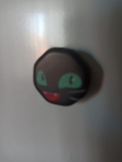 The image shows a black cat charm with green eyes and a red mouth, positioned on a light gray surface. The charm is blurred, suggesting it is either out of focus or in motion. The surface appears to be reflective, with areas of light and shadow. The scene is captured in low light conditions. There is no indication of a specific location in Promyshlennaya, Russia, or any details about people, actions, or interactions.