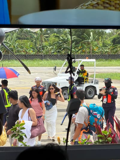 The image shows a group of people disembarking from a small airplane, possibly at Sayak Airport in Del Carmen, Philippines. Passengers are exiting onto the tarmac where a small vehicle is parked. Several ground crew members are present, assisting with luggage. One person holds a colorful umbrella. The environment includes tropical foliage and a paved area with markings, suggesting an airport runway.