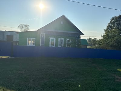The image shows a bright, sunny day in the village of Bolshie Shady, Russia. A traditional green wooden house with ornate white window frames stands in the center of the frame. A blue metal fence runs across the foreground, separating the house from a grassy yard. The sun is high in the sky, casting a bright glare on the scene. Trees and other buildings can be seen in the background. The overall impression is one of a peaceful rural setting.