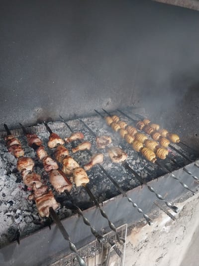This image shows a close-up view of food being cooked over coals on a grill. The primary subjects are skewers of food, including what appear to be spiral-cut potatoes and pieces of chicken. The scene is set outdoors, likely in a backyard or at a food stall, given the rustic nature of the grill and surrounding ground, which appears to be gravel and dirt. Smoke rises from the grill, indicating that the food is actively cooking. The lighting suggests it could be daytime, but there are no specific cues to determine the time of day precisely. The overall impression is one of preparing a meal, possibly a barbecue or a street food preparation. There is no visible text in the image.