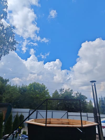 The image depicts an outdoor scene with a wooden hot tub in the foreground. The hot tub is octagonal with a dark exterior and a light-colored wooden interior. A metal frame extends upwards from the hot tub, and a stainless steel chimney pipe stands vertically to the right of it. In the background, a light-colored fence separates the yard from a line of trees. The sky above is a vibrant blue, dotted with fluffy white clouds. The overall impression is one of a backyard setting in a rural or suburban area, possibly in a dacha or country house environment. The location is identified as Лужники Снт, Russia. There are no people or specific actions evident in the scene.
