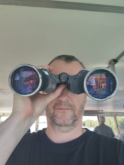 A man is depicted from the chest up, holding a pair of dark, slightly worn binoculars up to his face, gazing forward. He has graying hair and a short beard, wearing a black t-shirt with a gold chain around his neck. His expression is neutral and focused. Reflections in the binocular lenses show elements of the vessel's interior.

The setting is inside the cabin or bridge of a vessel, likely a boat, with a light-colored ceiling and structural beams. Through the windows in the background, a calm body of water, possibly a river or lake, is visible, lined with green, deciduous trees under a clear sky. Another man, with gray hair and wearing a gray t-shirt, is seen from behind in the background, looking out the windows, suggesting he is operating or overseeing the vessel's navigation.

The activity appears to be an observation or journey by water during daytime. The weather seems clear and bright. There is no visible text in the image. The scene is located in Chaika, Russia.
