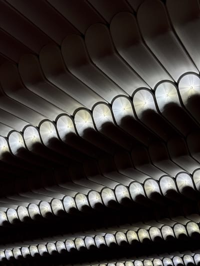 This media file is a close-up, abstract shot of a textured surface. The surface is composed of numerous overlapping, curved elements, possibly metal or a similar material, arranged in diagonal rows. Each element has a rounded end that tapers towards the point of overlap. Small, bright lights are embedded or shine from behind these elements, creating a dramatic effect of illumination and shadow. The lights are scattered across the surface, highlighting the texture and casting distinct circular reflections and soft shadows. The overall impression is one of a scaled or tiled pattern, possibly a facade or an architectural detail. The lighting suggests a low-light environment, perhaps nighttime, emphasizing the illuminated parts of the texture against the dark background. There is no visible text or human presence. The location context is Jermuk, Armenia, which might suggest this is an exterior architectural feature in that city.