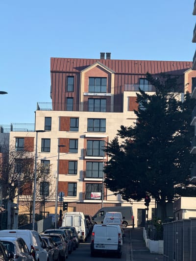 This image captures an urban street scene in Nogent-sur-Marne, France, under a clear, cloudless blue sky. A multi-story building, likely residential, occupies the background. Its facade is constructed with light beige stone or concrete panels interspersed with vertical reddish-brown wooden or metal panels. Numerous dark-framed windows are visible, many featuring black metal railings for balconies or Juliet balconies. The building's upper section includes several terraces with black metal railings and is capped by a sloped reddish-brown corrugated metal roof with multiple dormer structures and two dark chimneys. A red and white "A VENDRE" (For Sale) sign is affixed to a balcony railing on the building's lower-middle section.
In the foreground, a paved street runs from left to right. A row of vehicles is parked on the left side, consisting mainly of white vans and dark-colored passenger cars. On the right side of the street, a line of commercial vans, primarily white and grey, is stationary, indicating a halt in traffic, corroborated by a visible red traffic light. A large, dense evergreen tree with dark foliage partially obstructs the view of the central-right portion of the building and the right side of the street. No individuals are present in the scene. Street furniture includes a streetlight pole on the left and the traffic light pole on the right.