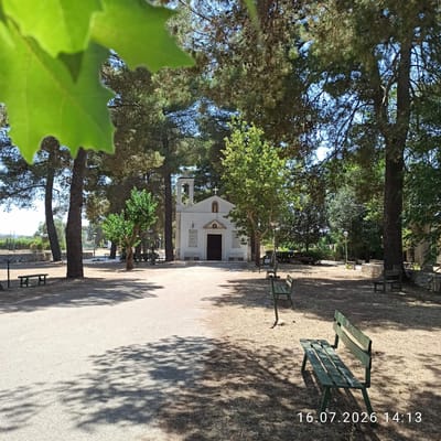 The image depicts a small white church with a bell tower and a cross on its facade, situated in a park-like setting with numerous trees. The church has a dark brown double door and two plaques on either side of the entrance. Several green benches are visible in the foreground and scattered throughout the park. The ground is a mix of dirt and dry grass, with shadows cast by the trees. In the background, there is a stone wall and more trees. The overall scene suggests a peaceful outdoor location. The timestamp in the lower right corner indicates the image was taken on July 16, 2026, at 14:13. While the image does not explicitly state its location, it is identified as being in Grumo Appula, Italy.