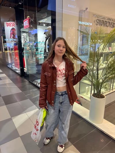 The image shows a young girl standing in what appears to be a shopping mall or retail environment. She is wearing a brown jacket, a white top, blue jeans, and sneakers. She is holding a strand of her hair in her hand and appears to be posing for the photo.
Behind her is a store window with various items on display, as well as a large sign that reads "SWAROVSKI." There is also a potted plant near the window. To the side, there is a sign with the text "DIESE" and "FOR SUCCESSFUL LIN" visible.
The flooring consists of gray and white square tiles. The scene is well-lit, suggesting an indoor environment.