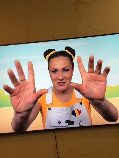 A woman with dark hair pulled into two buns is shown in a media file. She is wearing a bright orange shirt and white overalls with a colorful patch on the bib that resembles a house with a red roof. The background appears to be a cartoon landscape with a light blue sky and green and yellow hills. The woman has her hands held up towards the camera, palms facing outwards. Her facial expression suggests she is smiling and looking directly at the viewer. There is a ring on her left hand. The media file appears to be a screen capture or a video still, possibly from a children's educational program or entertainment show. The image is displayed on a screen, and the surrounding environment is visible, showing a textured brown surface that may be a wall or furniture. The lighting appears to be bright, suggesting an indoor setting with artificial light. No text is visible in the media file. The provided location context of Orhei, Moldova, does not offer any visual cues within the image itself.
