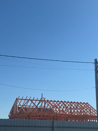 The image depicts a construction site in Matchino, Russia, under a clear blue sky. The central subject is the wooden framework of a house under construction, specifically its roof trusses, which are made of light-colored lumber and are arranged in a repeating pattern. The framework is partially covered by what appears to be dark roofing material. In the foreground, a grey metal fence runs horizontally across the bottom of the image, obscuring the lower portion of the construction. Overhead, three dark power lines stretch across the frame, and a utility pole with wiring is visible on the right side. No people or animals are present in the scene. The environment is open and appears to be a suburban or rural area, given the clear sky and the nature of the construction.