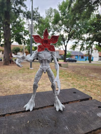A Demogorgon figure rests on a weathered wooden surface, possibly a picnic table, outdoors. The creature, characterized by its humanoid form and distinct flower-like head, is grayish-white. The floral head is red. In the background, there is a blurred landscape of greenery, trees, and a few buildings, indicative of a park or residential area.
