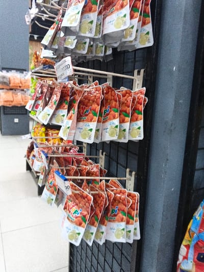 This image captures a retail store aisle, likely in Aglipay, Philippines, showcasing numerous small, individually packaged snack or candy products. The primary subjects are clear plastic pouches filled with reddish-orange, round items, resembling candies or dried fruits like rambutan or lychee. Each pouch features a white base with illustrations of a green/yellow fruit, possibly guava or lime.

The product brand appears to be "Yupi Delicious" as indicated by the visible text "pi" and "licious" on the packaging. Further details on the packaging include the words "Collagen" and "with real fruit juice," suggesting a health or fruit-based snack. A small logo is visible on the lower left corner of the white section of the packaging.

These pouches are neatly displayed on white metal racks with a black grid backing, characteristic of retail shelving designed for hanging products. There are multiple rows of these products, creating a repetitive pattern of vibrant packaging. A price tag in the middle section clearly displays "7.000."

In the background on the left, other blurred packaged items are visible on shelves, some in orange-hued bags, hinting at a variety of goods. The floor is light-colored and tiled. To the right, a dark gray, textured wall or pillar is seen, with a small portion of a colorful, patterned bag or textile in the bottom right corner. The lighting suggests an indoor, likely daytime, setting typical for a commercial establishment. No people are visible in the frame.