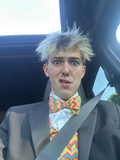 The image shows a young man in a black suit with a white shirt and colorful bow tie, sitting in the front seat of a car. The man has blonde hair with a spiky style and looks concerned with a slightly open mouth.  He is wearing a black suit jacket, a white shirt, a colorful patterned bow tie, and a matching colorful patterned waistcoat.  He is wearing a black seatbelt, which is fastened across his chest. The car's interior is visible in the background.  The sky is blue, and trees can be seen through the rear window of the car.  The image was taken inside a vehicle in the city of Minskip, UK (latitude: 54.066, longitude: -1.39), which is located in North Yorkshire.
