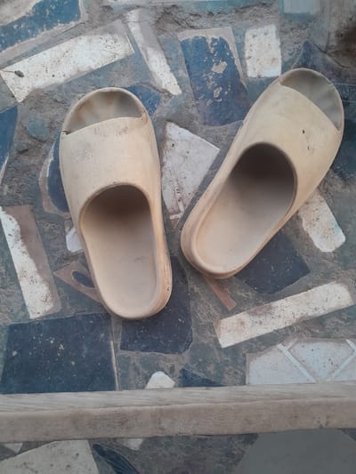 This image depicts two beige slide sandals resting on a patterned tiled ground in Ifo, Nigeria. The sandal on the left is oriented vertically, toe pointing towards the top of the frame. The sandal on the right is diagonally oriented, with its toe pointing towards the upper right. Both sandals show signs of wear, including dirt accumulation and minor surface abrasions. The ground consists of an arrangement of irregularly shaped tiles in various shades of dark blue, grey, and off-white, also exhibiting dirt and wear. A weathered, light brown wooden plank or beam extends horizontally across the lower portion of the image. No individuals, actions, or interactions are present in the scene.