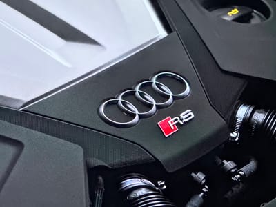 The image shows a close-up view of an Audi RS engine. The iconic four rings logo is prominently displayed, along with the "RS" badge in red. The engine cover is dark and textured, with some metallic elements visible. The background is dark and out of focus, suggesting an engine bay environment. There are no people or any identifiable features of the city of Bezden, Bulgaria in the image.