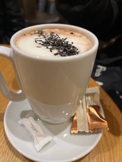 An close-up, indoor image depicts a white ceramic mug containing a hot beverage, likely hot chocolate or cappuccino, topped with a layer of white foam and numerous dark brown chocolate sprinkles. The mug is positioned centrally on a white ceramic saucer, which rests on a light-colored wooden surface, presumably a table. To the left of the mug on the saucer is a single, individually wrapped white sachet, possibly containing sugar or artificial sweetener. To the right of the mug, also on the saucer, lies a single, individually wrapped golden-brown packet, consistent with a biscuit or small chocolate bar. The background is blurred and dark, with a faint, partial white text reading "Fibo" or similar visible on the right side. The environment suggests a café or restaurant setting in the city of Sélestat, France. No individuals are explicitly visible.