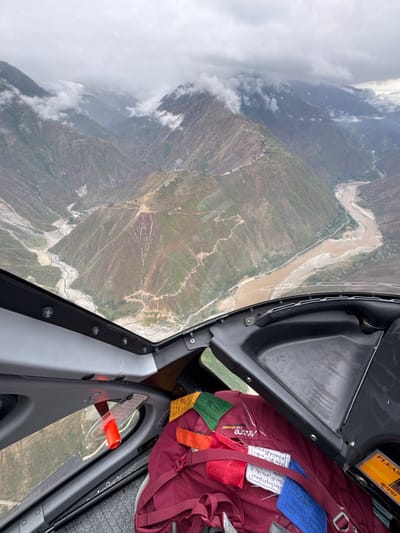 This media file captures an aerial view from inside an aircraft, looking out over a rugged mountainous landscape. The perspective is high-angle, revealing vast valleys and peaks.

In the foreground, part of the aircraft's interior is visible, including a gray and black window frame on the right. A red trekking backpack is stowed beside the window, adorned with colorful Tibetan Buddhist prayer flags in blue, red, and green. Text visible on the backpack includes "Mirzone" and "Trek ND43:50". Above the backpack, a yellow sign partially reads "PLEAS" and "कृपया" (Kripaya, meaning "Please" in Hindi/Nepali). A small red handle or lever is also visible near the window.

The expansive scene outside the window features a wide, muddy-brown river meandering through a deep valley. The riverbanks and surrounding slopes show signs of small settlements and agricultural terraces, connected by winding dirt paths. The mountains appear steep and largely barren, with patches of green vegetation. The sky is overcast, with clouds clinging to the mountain peaks and scattered throughout the valleys, suggesting a cloudy but daytime weather condition.

The overall scene depicts travel over a remote, high-altitude region, consistent with the cultural cues of the prayer flags and the provided location context of Jinakhu, Nepal. The image evokes a sense of journey and the dramatic scale of the natural environment.