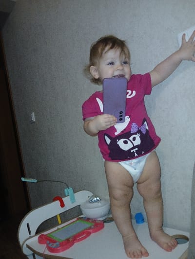 A baby lies on her back, holding a purple phone to her face. She wears a pink shirt with a cat graphic and a diaper. Her legs are extended, one foot slightly elevated. A blue bottle cap rests near her legs. A white and red rocking horse toy with a drawing pad is in the background. The background also includes a white surface, possibly a crib or play area. The floor is a light gray, neutral carpet.