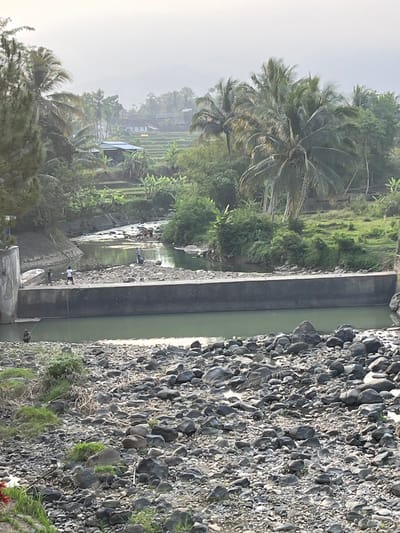 This image captures a serene, natural landscape in Krasak, Indonesia. The scene is dominated by a river flowing through a rocky terrain. A concrete dam, partially submerged, spans across the river, creating a calm pool on one side and a more turbulent flow on the other. Several individuals are present, engaged in various activities. One person is fishing from the rocky bank, holding a fishing rod. Others are standing on the dam, seemingly observing the water or interacting with each other. The surrounding environment is lush and green, with dense vegetation, palm trees, and distant terraced rice fields adding to the picturesque setting. The sky is overcast, suggesting a hazy or early morning light.