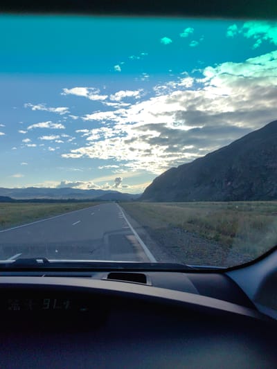 An asphalt road stretches into the distance, bordered by dry grass and a large mountain on the right. The sky is partly cloudy, with a bright sun peeking through. The image is taken from inside a car, with a dashboard visible in the foreground displaying a time of 9:11. The location is identified as Boyarovka, Russia.