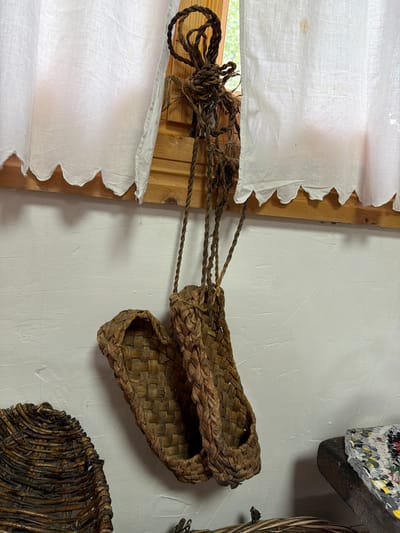 A pair of woven, natural fiber sandals hangs by their braided rope straps from a wooden support beam in front of a white, textured wall and a white linen curtain with a scalloped hem. The sandals are made of what appears to be straw or reeds, woven in a basket-like pattern, with a braided edge and a toe strap. They are positioned in the center of the frame, slightly angled. To the left, the edge of another woven basket is visible. To the right, a dark, rough-hewn wooden object supports a colorful woven mat or textile. The lighting suggests an indoor setting, possibly a rustic dwelling or museum exhibit, with natural light filtering in from a window behind the curtain. The overall impression is of handcrafted, traditional footwear displayed in a simple, unadorned environment.