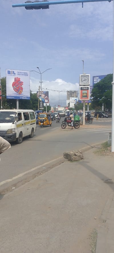 This image depicts a bustling urban street scene in Tudor ward, Kenya, viewed from a low angle. The sky is light blue with scattered white cumulus clouds.
In the immediate foreground, to the lower left, a partial view of a person's light-colored clothing is visible. The foreground road surface is grey asphalt with some minor cracks and debris. A concrete curb separates the main road from a dirt and grass verge on the right, where a large, irregular grey rock rests.
The midground features a multi-lane asphalt road with vehicular traffic. Dominantly, a white Nissan Caravan/Urvan van with yellow stripes is positioned on the left, facing away from the viewer. To its right, a yellow auto-rickshaw (tuk-tuk) is visible. Numerous motorcycles (boda-bodas) are present; one motorcyclist, wearing a white helmet and dark clothing, is stationary in the center-right lane, with a green delivery box attached to the rear of his motorcycle. Other cars, vans, and trucks are discernible further down the road, indicating a busy traffic flow. People are visible as drivers and passengers within the vehicles.
The background consists of various urban elements. On the left, a large billboard for "ASL Wires & Cables" displays "THE PULSE BEHIND EVERY CONNECTION" alongside an illustration of a human heart formed by colorful wires. A streetlight pole stands nearby. To the center-right, a tall pole supports a square clock showing "CITY" and "Mombasa" (partially visible), with an analog display. Below the clock, another square advertisement for "MIKA" appliances (depicting a rice cooker) is affixed to the same pole. Additional billboards further down the street advertise "MIKA" again and "LOVE MILK AGAIN." Multi-story buildings, including one under construction with exposed rebar and concrete, line the street in the distance. Green foliage from trees is visible behind some billboards. Overhead, a light blue metal gantry structure spans the road, holding what appear to be traffic lights.