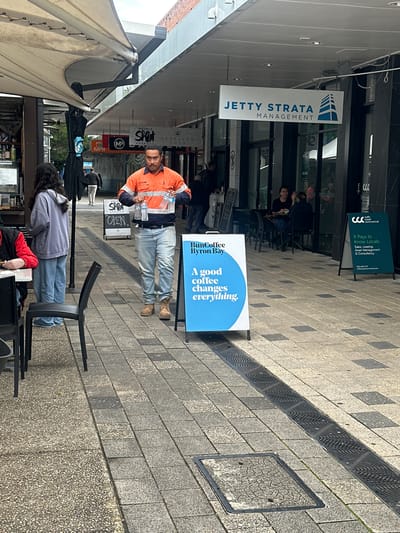 The image depicts a street scene in Coffs Harbour, Australia. A man in an orange high-visibility work shirt and blue jeans is holding multiple bottles of water while walking. He is positioned in the center of the frame, on a paved sidewalk. To his left, a woman in a grey hoodie and blue jeans stands near a cafe. Several tables and chairs are visible on the sidewalk, suggesting an outdoor dining area. Various signs are present, including one for "Jetty Strata Management" and another advertising "Bun Coffee Byron Bay" with the slogan "A good coffee changes everything." A sign for "Coff's Coast Commercial" is also visible. The environment appears to be a commercial street with shops and a canopy overhead. The lighting suggests it is daytime, with overcast skies. The pavement is a mix of stone tiles and aggregate.