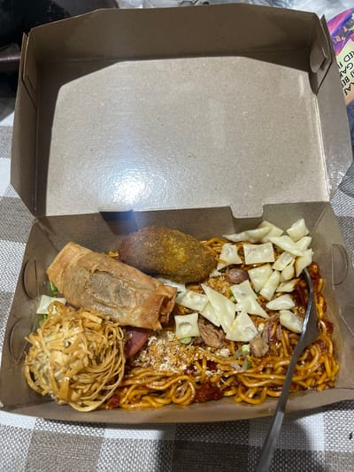 A top-down, close-up shot shows a cardboard takeout box filled with noodles and various side dishes. The box is open, revealing the contents arranged in the right half of the container. The left half of the box is empty and casts a shadow from an unseen light source. The background is a checkered tablecloth in shades of grey and white.

The primary subject is a serving of savory noodles, tossed in a reddish-brown sauce, with some visible scallions and shredded fried garlic scattered on top. Interspersed within the noodles are pieces of what appear to be sausage and several light-colored, diced ingredients, possibly wonton skins or tofu. A metal fork rests on top of the noodles, partially submerged.

To the left of the noodles are three distinct side dishes. The uppermost is a golden-brown, irregularly shaped fritter or croquette. Below that is a larger, rectangular item wrapped in a thin, crispy pastry, resembling a spring roll or a similar fried snack. To the bottom right of these is a mound of crispy, golden-brown noodles, coiled into a nest-like shape and drizzled with a light sauce.

A portion of text is visible on the lid of the takeout box, to the upper left of the frame. It is partially obscured and appears to be in an Asian script, possibly Indonesian, with some English words like "RED F" visible. The lighting suggests it may be indoors or evening. The overall scene depicts a meal ready for consumption, likely prepared for takeout. The setting is casual and domestic, indicated by the tablecloth.