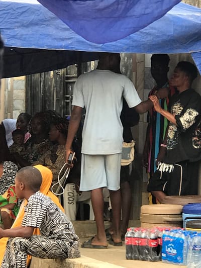 Several people are gathered under a makeshift structure, with a blue tarp visible on the left. Some individuals are standing, while others are seated on the ground. A man in a light blue shirt is looking at a phone. Bottled water and other items are stacked on a shelf. People are wearing casual clothing, and the overall scene suggests a gathering or waiting area.