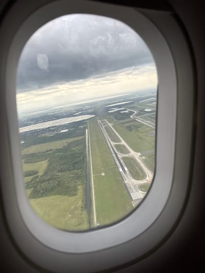 The image captures an aerial view from an airplane window, looking down at an airport runway and surrounding landscape. The dark, curved frame of the airplane window is prominent in the foreground, framing the scene outside.
The view outside is significantly tilted, suggesting the aircraft is banking during takeoff, landing, or a low-altitude maneuver. On the right side of the frame, a large section of an airport runway system is visible. The main runway is clearly marked with white lines, including threshold stripes and the number "17." Several paved taxiways curve and connect to the main runway, all surrounded by extensive areas of vibrant green grass.
Further into the landscape, a distinct body of water, possibly a canal or river, runs parallel to portions of the airport infrastructure. Beyond this, the terrain features patches of dense green trees and more open fields. The sky on the left side of the window appears overcast, with varying shades of gray and white clouds. The lighting indicates it is daytime, possibly under partly cloudy conditions. Given the provided context, this scene depicts an airport in Orlando, United States of America.