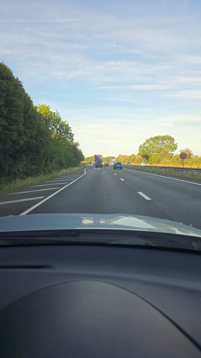 The image depicts a view from the driver's perspective of a car on a highway. The highway has multiple lanes, indicated by white lines. On the left side of the highway, there is a dense line of trees. On the right side, there are trees and a guardrail with a red circular sign. Several vehicles are visible on the highway, including a blue car in the middle lane, a blue truck in the far lane, and other cars and trucks further ahead. The sky is blue with scattered white clouds. The foreground shows the dashboard of the car. The scene suggests a typical daytime drive on a UK highway, potentially within Cherwell District, United Kingdom, given the presence of characteristic highway infrastructure and vegetation.