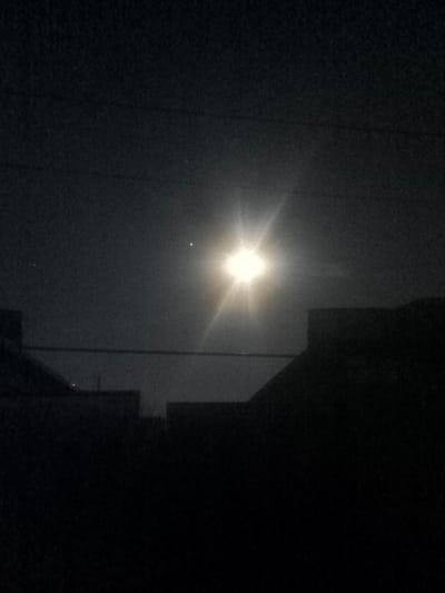 This image depicts a nighttime scene observed in Pakhtakor, Uzbekistan. The upper portion of the frame is dominated by a dark, grainy sky. A single, intensely bright celestial body, likely the Moon, is positioned slightly right of center, exhibiting a prominent starburst effect. To the left of this primary light source, a smaller, distinct white point of light is visible, possibly a planet or star. Further to the left, a fainter, smaller white point of light is also present. A thin, dark horizontal line, consistent with an overhead utility wire or cable, traverses the middle ground of the image, running below the primary light source. The lower portion of the image is occupied by the dark, indistinguishable silhouettes of multiple building rooftops or structures, characterized by angular outlines against the subtly illuminated sky. No people, discernible actions, or interactions are visible.