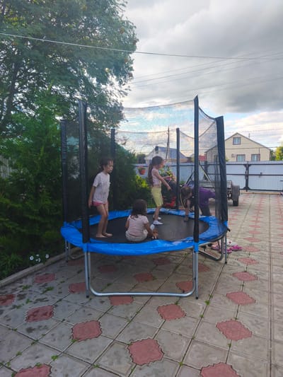 In Burayevo, Russia, a blue trampoline with a safety net is set up on a patterned tiled patio. Four children are on the trampoline, engaged in various activities. One child, a girl with long dark hair, stands with her legs apart, looking to the side. Another child, a girl with lighter hair, sits facing away from the camera. Two other children are in the process of interacting with each other on the trampoline. To the left of the trampoline is lush green foliage, and to the right, a light-colored building with a pitched roof is visible, along with a white fence and a trailer. The sky is partly cloudy, with some visible power lines overhead.