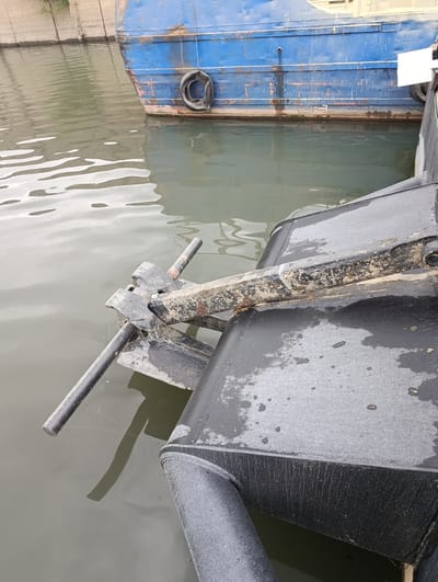 The image captures a close-up view of a blue barge docked in a body of water, likely a canal or river, in Šabac, Serbia. The barge's hull shows signs of wear, with peeling paint and rust streaks visible. A black tire is attached to the side of the barge, serving as a fender. In the foreground, a black mechanical component, possibly part of the barge's equipment or a separate structure, is partially submerged in the murky water. The water's surface is rippled, reflecting the overcast sky. The environment appears industrial, with concrete structures visible in the background. The scene is devoid of human presence.