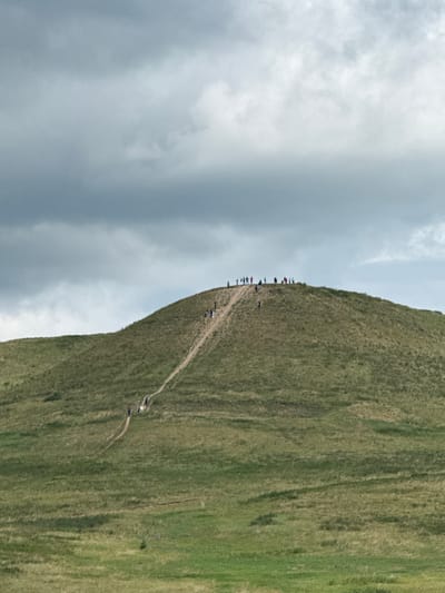 The image depicts a grassy hill with a winding dirt path leading to its summit. A group of people are scattered along the path and at the top, appearing to be hiking or exploring. The sky above is overcast with dramatic, dark clouds, suggesting a moody or potentially stormy atmosphere. The foreground consists of more rolling, green terrain. The overall impression is of an outdoor recreational activity in a natural, somewhat rugged landscape. The location is stated to be in the city of Nuryevo, Russia.