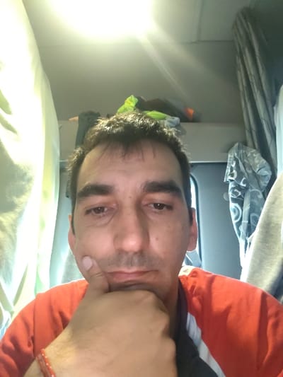 The image displays a male individual, appearing middle-aged, with short, dark, somewhat disheveled hair, dark eyebrows, and brown eyes. His gaze is directed downwards or appears heavy-lidded. He is positioned in a head-and-shoulders view, with his right hand raised, thumb supporting his chin, and fingers curled. He wears a red and dark-colored shirt, and a red beaded bracelet is visible on his right wrist. The background features the interior of a vehicle cabin, consistent with a truck's sleeper compartment. Above the subject, an overhead shelf or bunk is visible, containing various items including a bright yellow/lime green fabric item and darker clothing. A strong light source is present in the upper center, creating a glare. Curtains frame the scene; a light-colored, possibly white or pale green, curtain is on the left, and a grey patterned curtain, pulled back to reveal a window or door frame, is on the right. No specific visual markers in the image allow for the identification of the city of Servas, France, or a precise location within it.