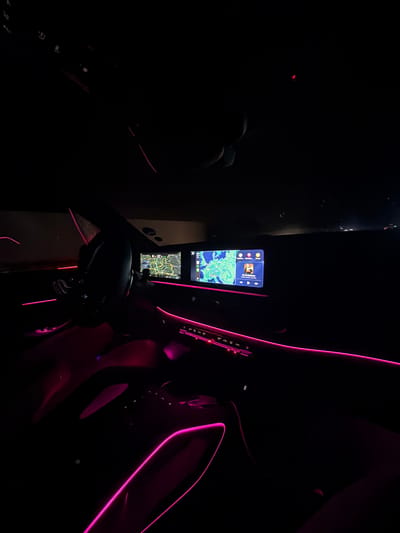 The image shows the interior of a modern car at night, illuminated by pink ambient lighting. Two screens are visible on the dashboard; the left screen displays a map of Europe, while the right screen shows album art for a song titled "Clip To The Horizon" by Corinne Bailey Rae, along with playback controls. The steering wheel is visible in the foreground, and the car's interior appears spacious and luxurious. The scene is dark, with only the screens and ambient lighting providing illumination. The location is identified as the city of Plauen, Germany.