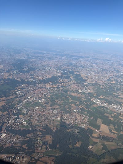 This is an aerial photograph of the city of Vizzola Ticino, Italy, taken from an airplane. The image shows a sprawling urban landscape interspersed with agricultural fields and patches of forest. The city itself appears to be densely populated with buildings of various sizes and colors, forming a mosaic of residential and commercial areas. Roads and infrastructure are visible, connecting different parts of the city and extending into the surrounding countryside. The agricultural land is divided into rectangular plots, some of which appear to be cultivated with crops, while others are fallow or covered in vegetation. Patches of dark green forest dot the landscape, providing a contrast to the more developed areas. The sky above is a clear blue, with a few wispy clouds visible in the distance. The overall impression is one of a vibrant, active community integrated into its natural surroundings.