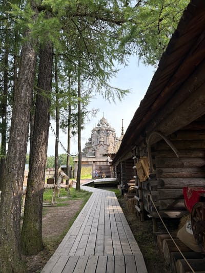 The image depicts a wooden walkway leading towards a traditional Russian wooden church with multiple domes, set against a bright, overcast sky. The walkway is constructed from light-colored wooden planks and is angled from the lower right to the upper left of the frame. To the left of the walkway, a row of mature pine trees with rough bark and green needles lines the path. The background shows more wooden structures, possibly part of an open-air museum or historical park, with a hint of green grass and foliage.

In the distance, the elaborate wooden church structure is the focal point, its intricate carvings and multiple cupolas suggesting a historical or religious significance. The presence of string lights with white bulbs, strung between trees, suggests the area might be used for events or that it is nearing evening. A few small figures of people can be seen near the entrance to the church, giving a sense of scale to the impressive architecture.

The overall scene evokes a sense of cultural heritage and natural beauty, characteristic of a park or historical site in Russia. The lighting suggests it is daytime, with diffused light indicating overcast conditions, likely making for a mild day. The rustic architecture and natural setting are dominant themes.